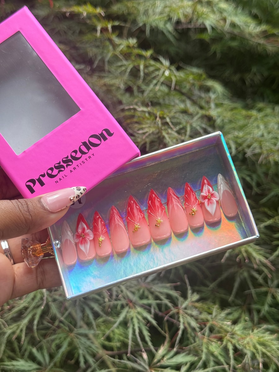 Set of medium length stiletto press-on nails featuring a refined red French tip design, gold charms, and a delicate 3D flower accent.