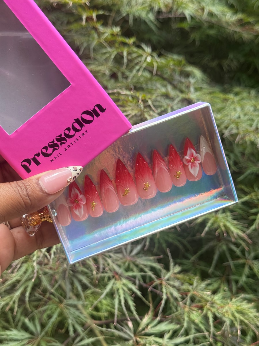 Medium almond shaped press-on nails with a pink and red ombre design. Some nails have small white flower decorations and tiny gold accents.