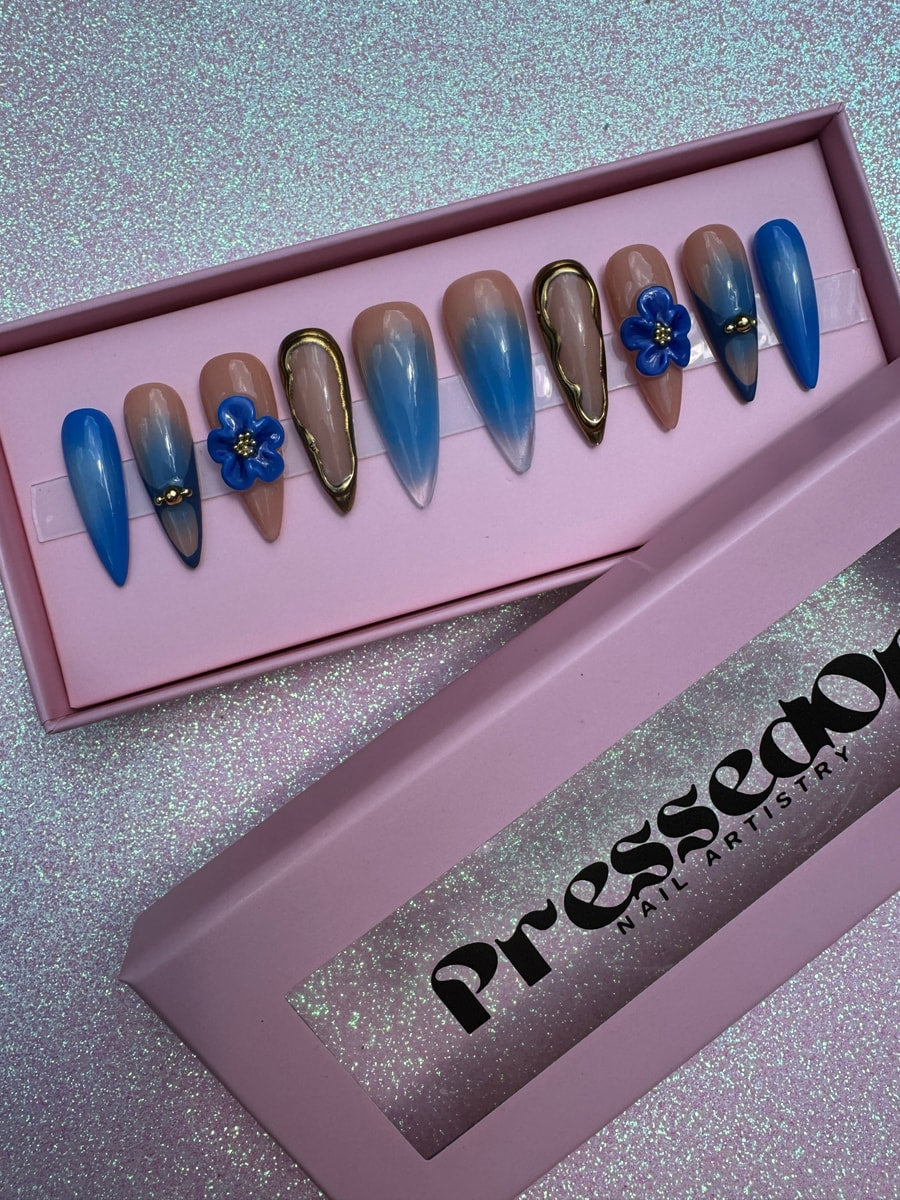 Royal blue medium press-on nails include a nude ombre, gold outlines, and blue 3D flower accents. The nails are stiletto and rounded shapes.