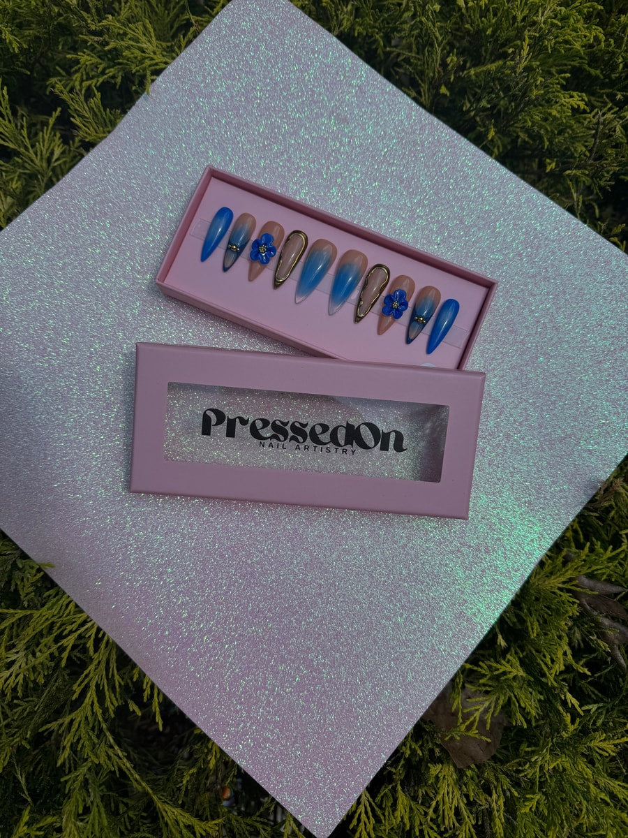 Royal blue medium-length press-on nails feature a nude base fading into a blue ombre tip, adorned with gold trim and small blue flower accents. The design alternates between the two styles.