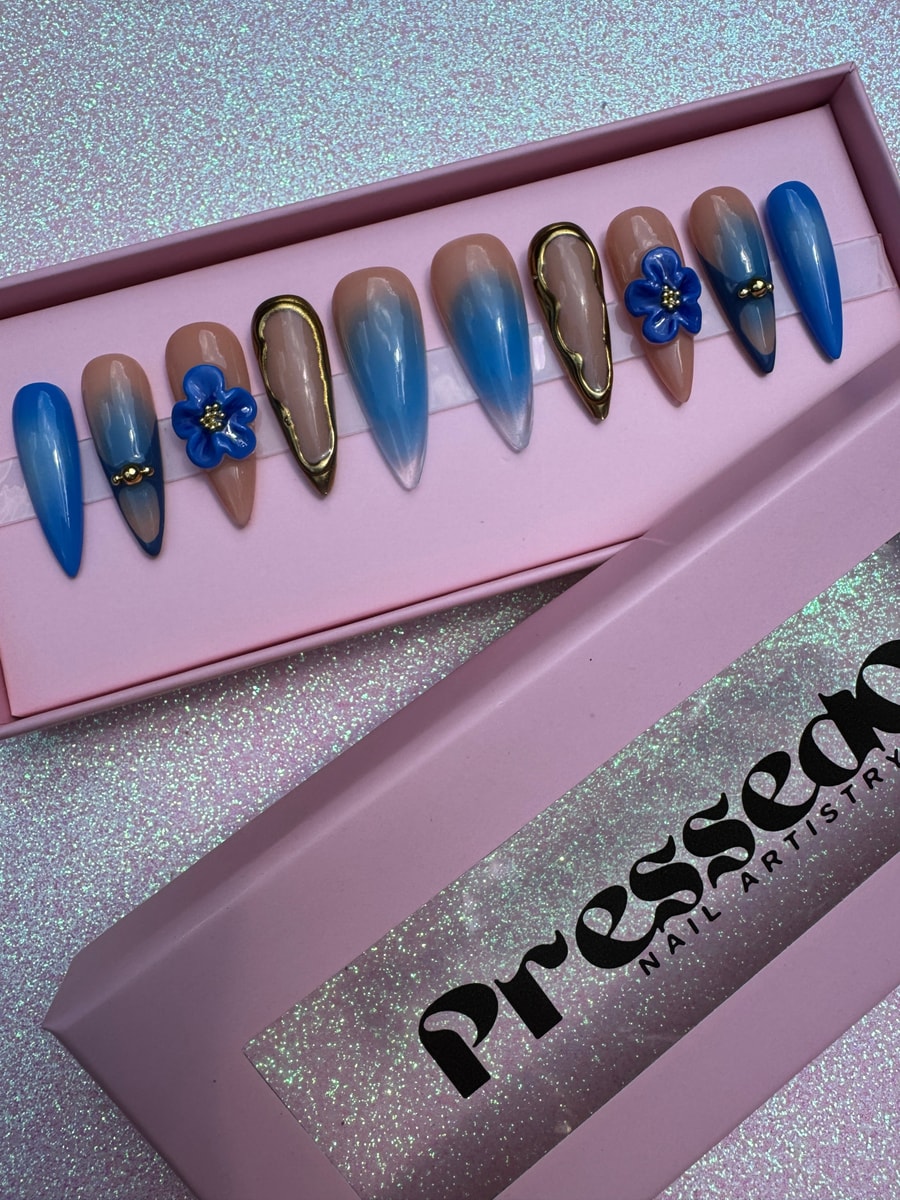 A set of medium press-on nails features royal blue ombre designs, gold accents, and blue flower charms on a nude base.