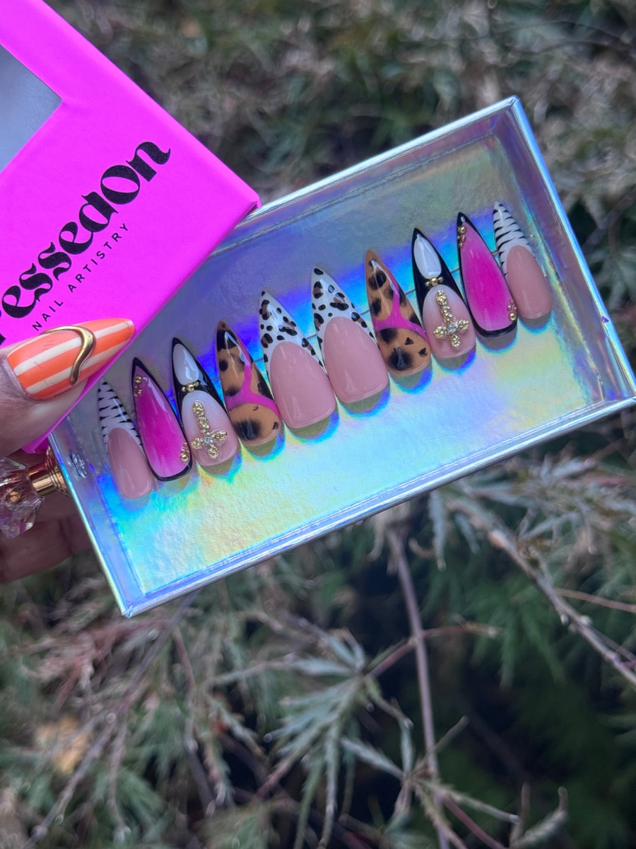 Medium-length press-on nails feature a safari chic design with leopard print, zebra stripes, Dalmatian spots, solid pink nails, and gold cross charms.