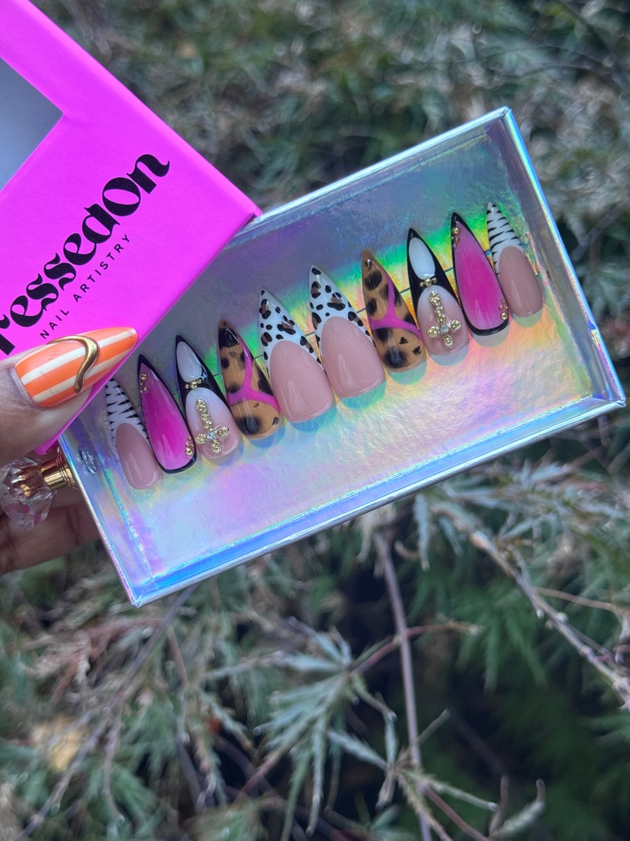 Press-on nails with a safari chic theme. The nails feature various animal prints on the tips, including leopard, cow print, and zebra stripes. Some nails have pink accents and small gold cross charms.
