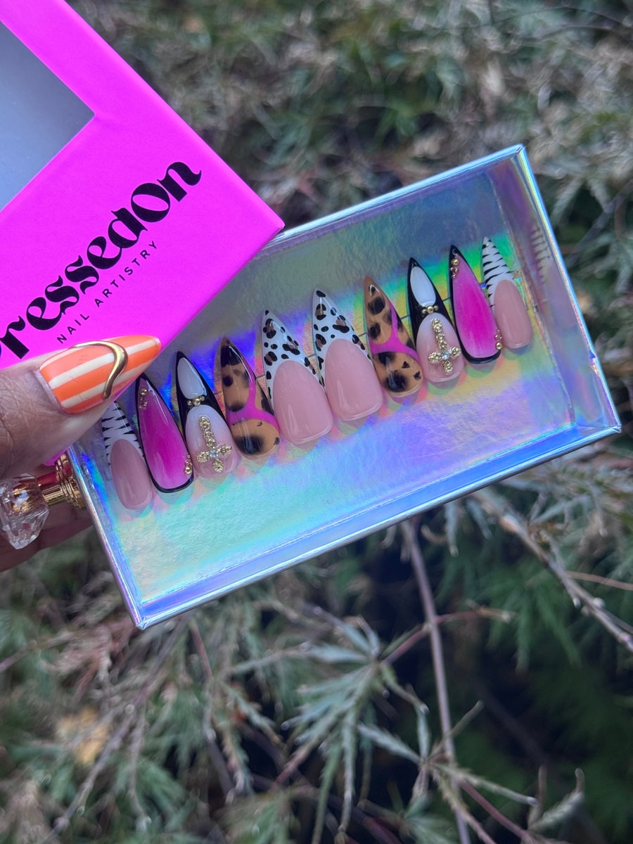 Stiletto press-on nails with safari-inspired designs, including leopard and zebra prints, pink ombre, and nude nails. Gold crosses and studs adorn some nails.