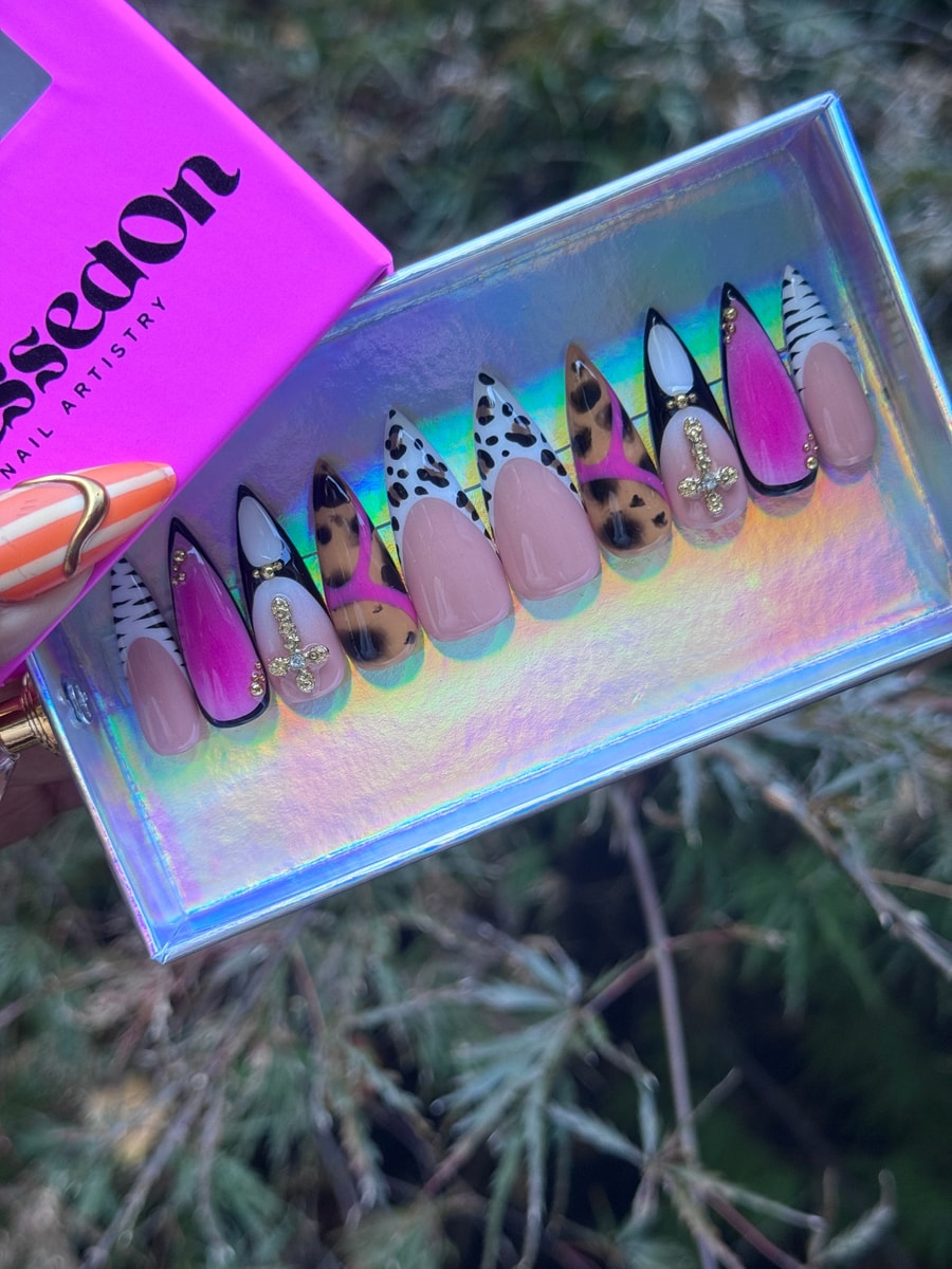 The image showcases a set of press-on nails featuring a safari theme. The nails have animal print designs like leopard spots and zebra stripes, along with shades of pink and gold cross charms.