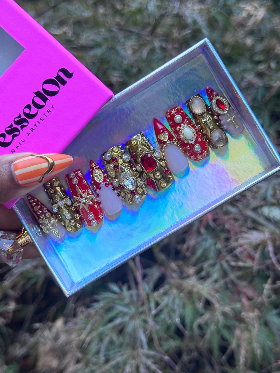 Ornate red and gold press-on nails are adorned with pearls, jewels, and crosses. Several have a solid red or white base.