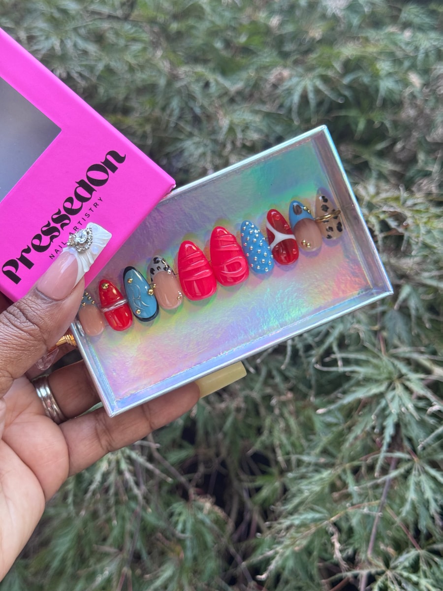 This image shows short almond press-on nails. They feature red, blue, and neutral designs with gold accents and leopard print details.