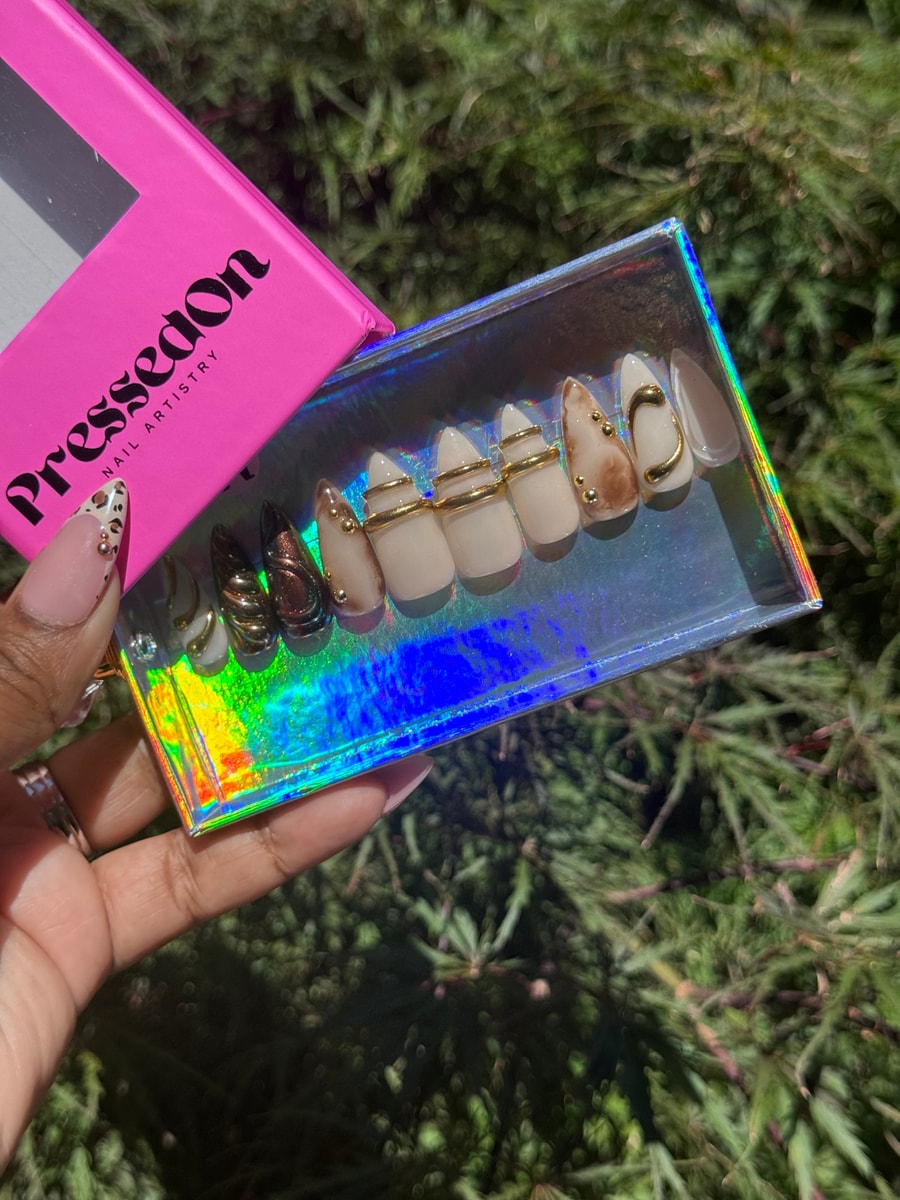 Short, almond-shaped press-on nails with a chrome and neutral design. Some nails feature gold ring and swirl accents and scattered gold embellishments.