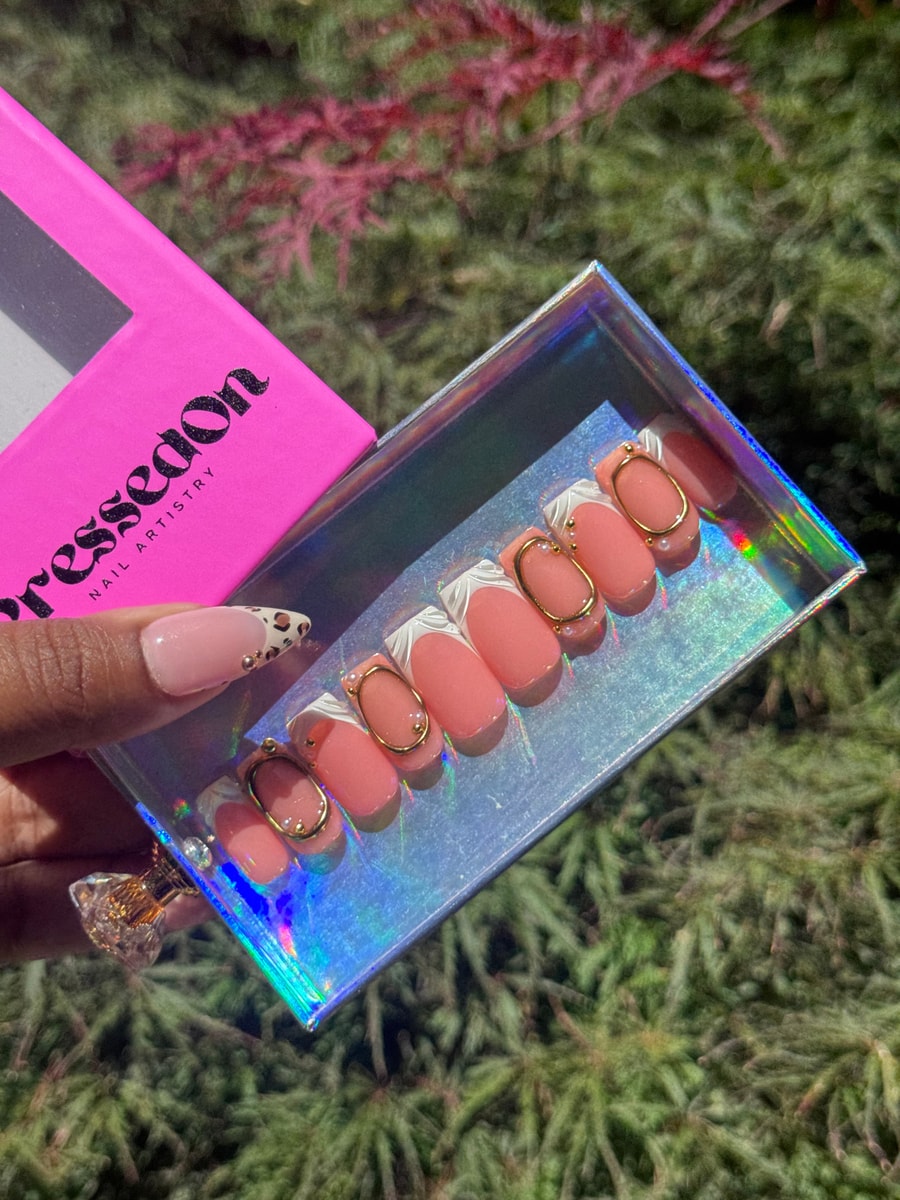 Peach-colored short press-on nails featuring a white French tip and unique 3D gold oval accents. One nail is adorned with a nude and leopard print design accented with small gold gems.