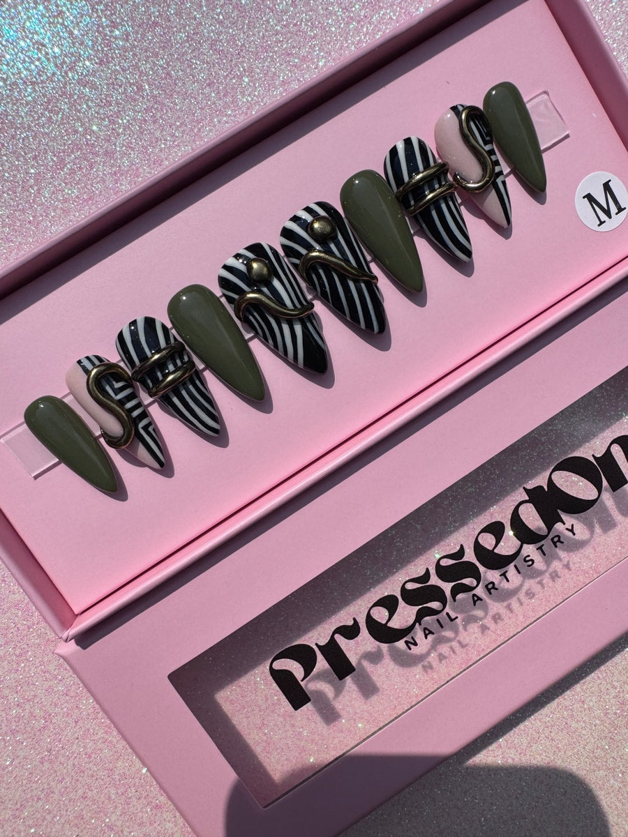 Short stiletto press-on nails with a geometric design featuring olive green, black and white stripes, and a blush pink accent nail. Some nails have a gold embellishment.