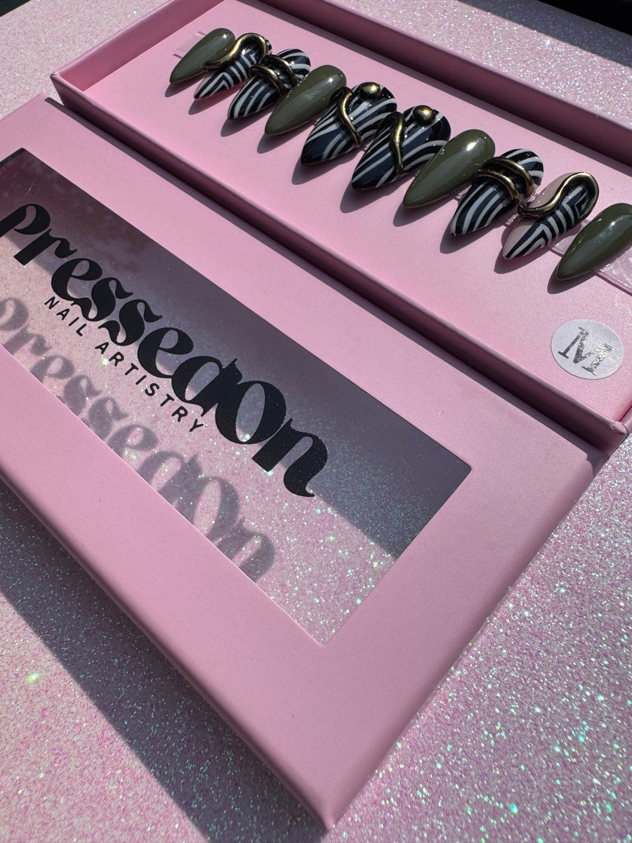 Short press-on nails feature a mix of zebra stripes and olive green designs. Some nails have a gold metal accent.