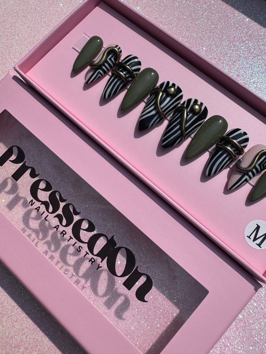 Short geometric press-on nails with zebra stripes in black and white, solid olive green nails, and gold-toned metal decorative elements. 