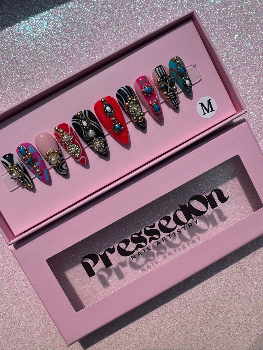 A set of short, stiletto press-on nails featuring varied designs including black and white patterns, pink, teal, and red, each embellished with decorative charms.