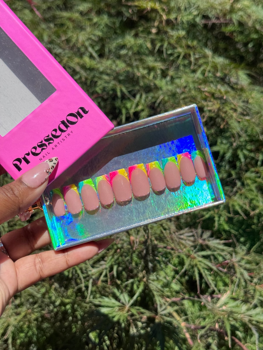 Short nude press-on nails feature a colorful neon French tip design in pink, yellow, green, and blue.