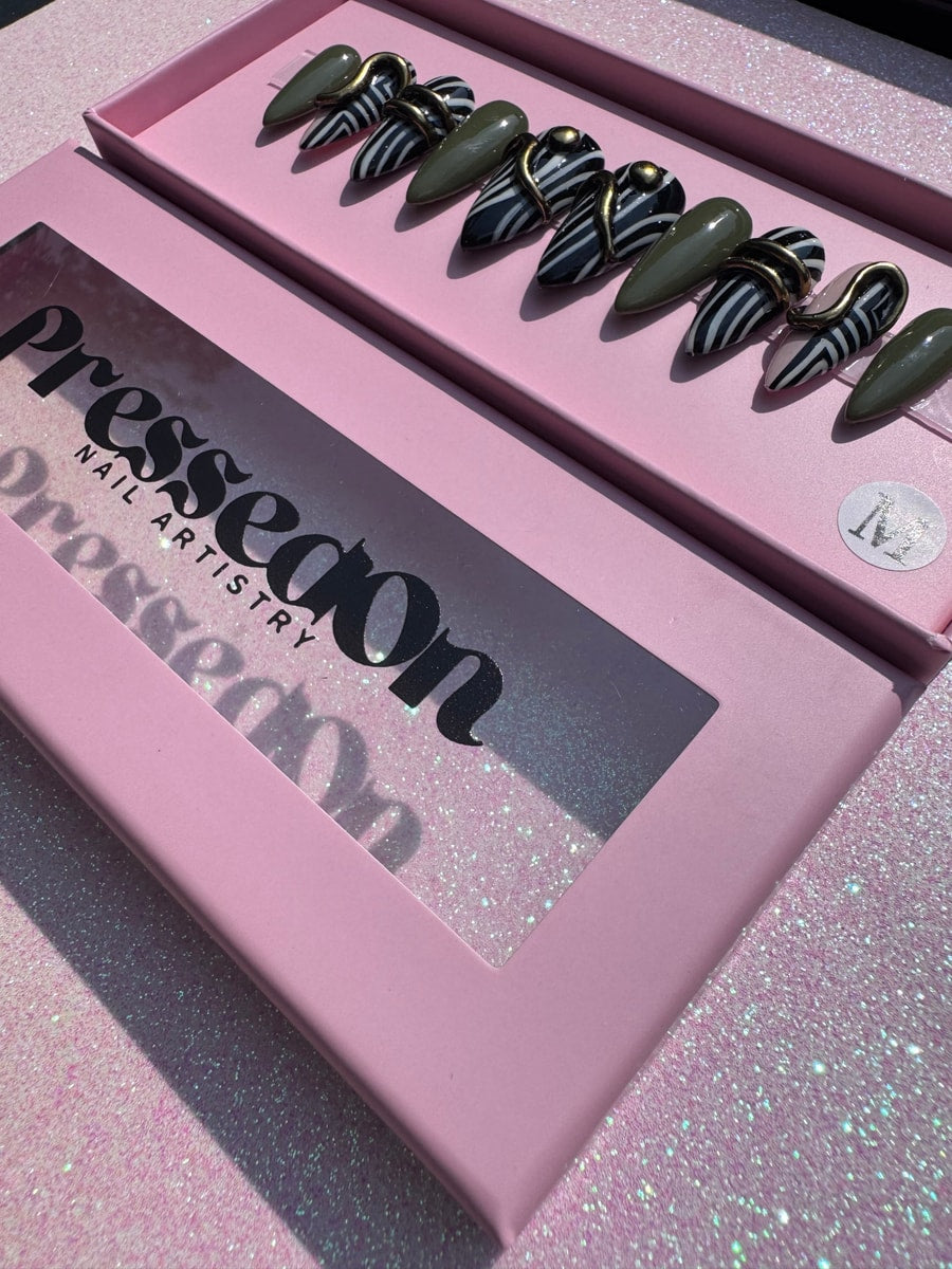 A set of short, stiletto-shaped press-on nails includes olive green nails and zebra-striped nails, some decorated with gold bands or studs.