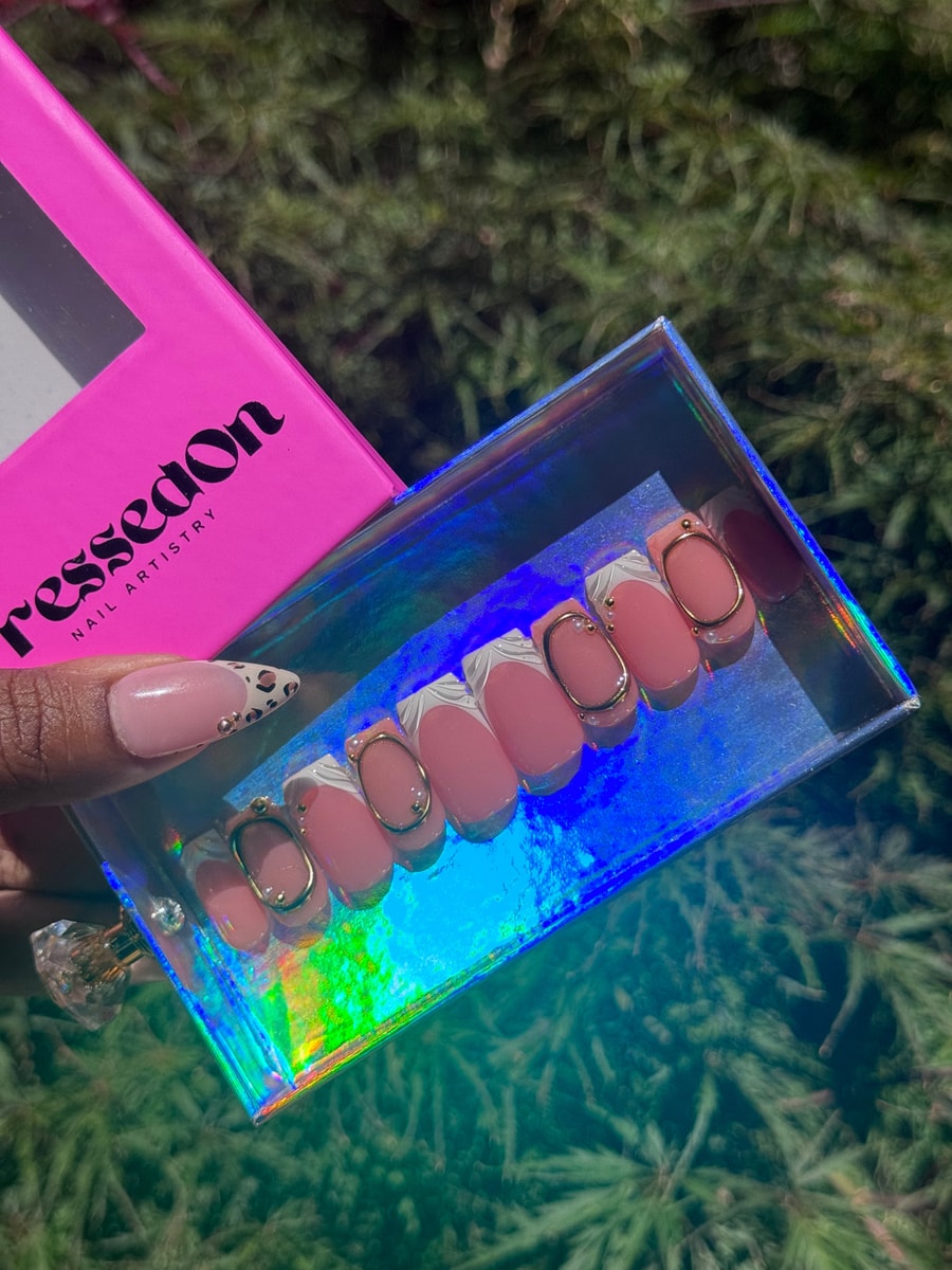 Short pink press-on nails with white French tips and gold oval accents. One nail has a pink base with a leopard print tip.