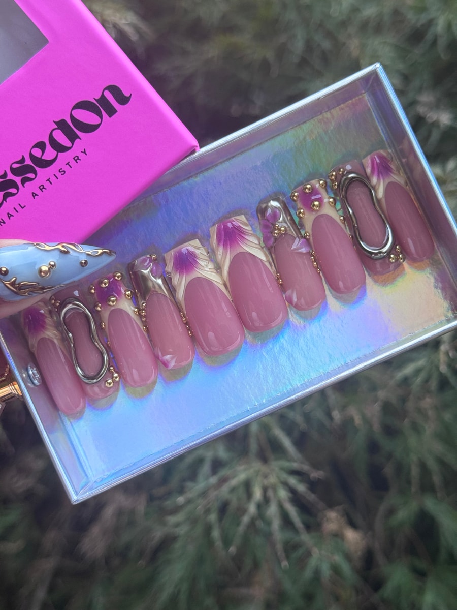 Short pink press-on nails featuring a white French tip with purple floral accents and gold bead embellishments. Some nails also have silver hardware.