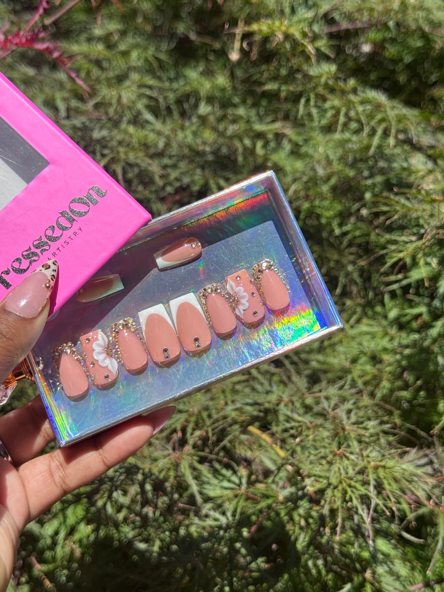 A set of short pink press-on nails with rhinestone and flower accents. The set includes French tip styles and some with rhinestones.
