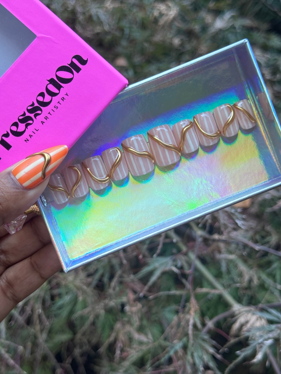 Short squoval press-on nails with a pink and white striped pattern and a curvy gold accent on each nail. One orange striped nail is included.