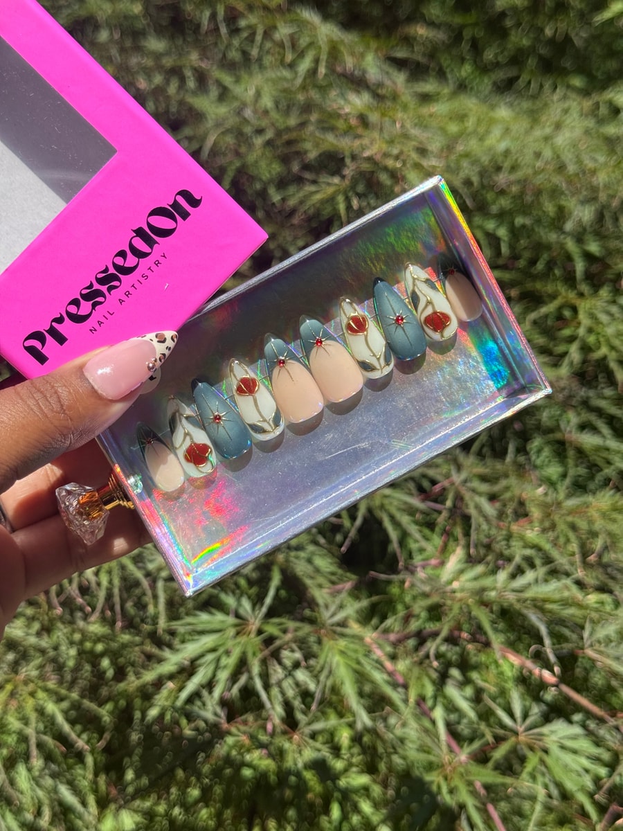 A set of short press-on nails featuring a Tea Time floral design with red and gold accents on a mix of neutral and blue-gray nails. Small red gems adorn some of the nails.