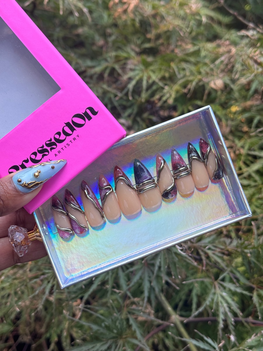 These are short stiletto press-on nails with nude bases, darker color accents at the tips, and silver wire decorations. One nail is light blue with golden details.