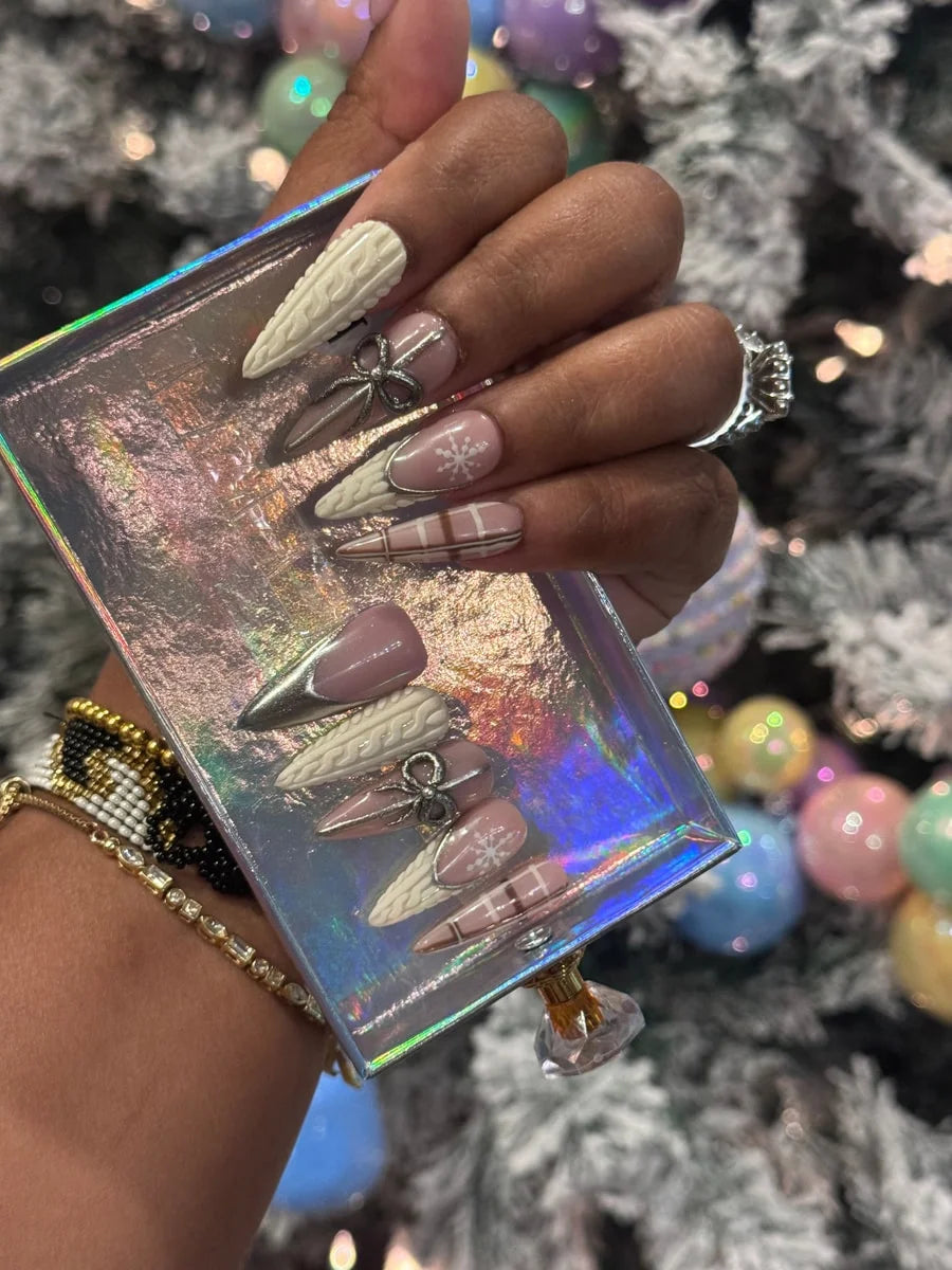 Medium-length press-on nails with cashmere-inspired designs including cable knit textures, plaid patterns, snowflakes and silver bow embellishments on a set of nude pink, metallic silver and cream colored nails.