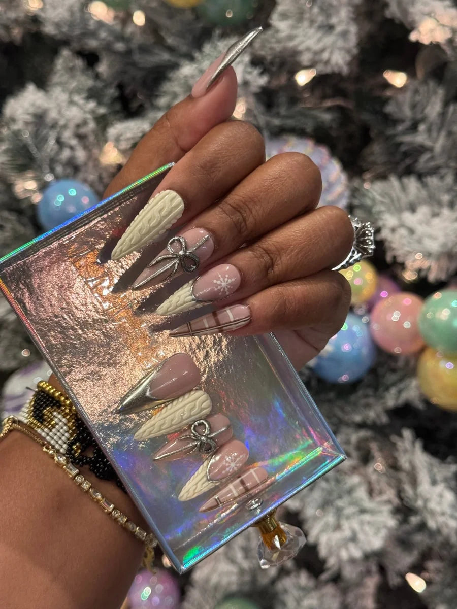 Medium-length press-on nails featuring a cashmere-inspired design, including cable knit textures, plaid patterns, a snowflake design, and silver bow embellishments, with nude pink and metallic silver accents.