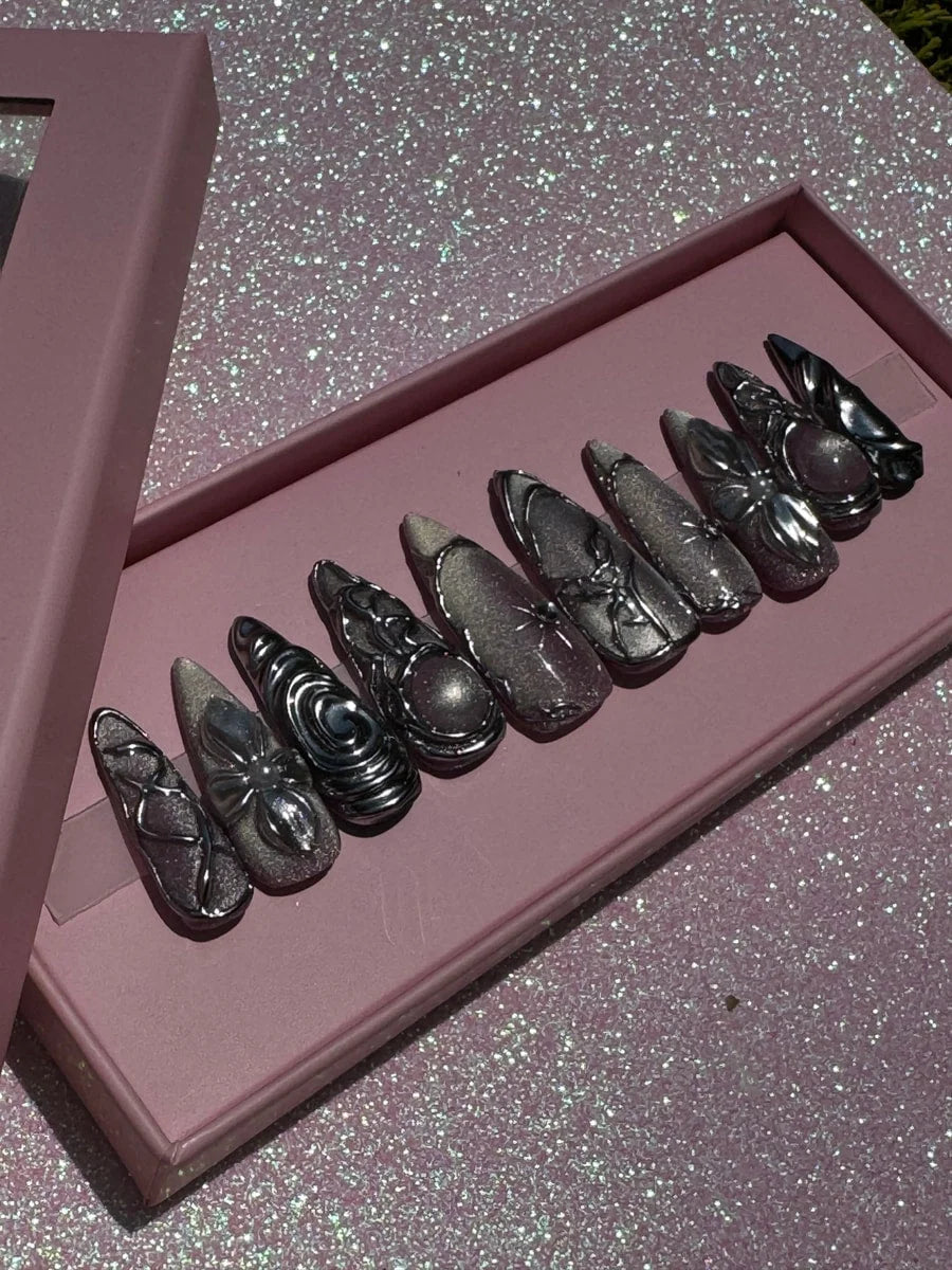 Medium length chrome press-on nails featuring glitter details and ornate 3D silver charms. The nails include swirling designs and pink stone accents.