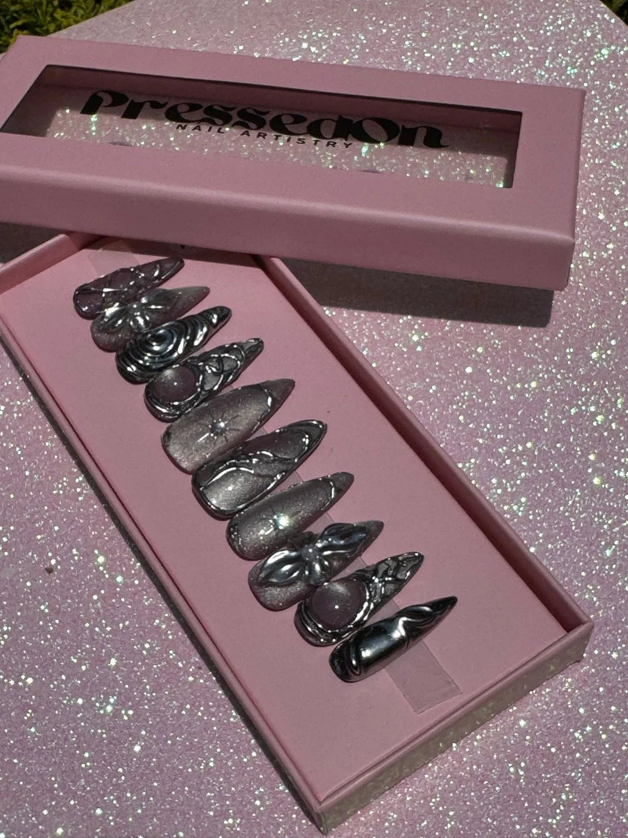 Medium-length chrome press-on nails featuring 3D silver charms and glitter accents. Some nails have pink stone accents.