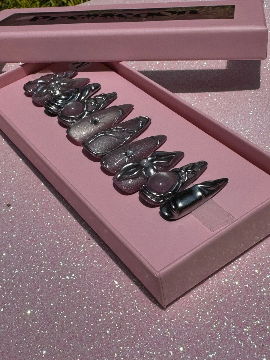 Medium-length chrome press-on nails featuring 3D silver charms, glitter accents, and pink stone details.