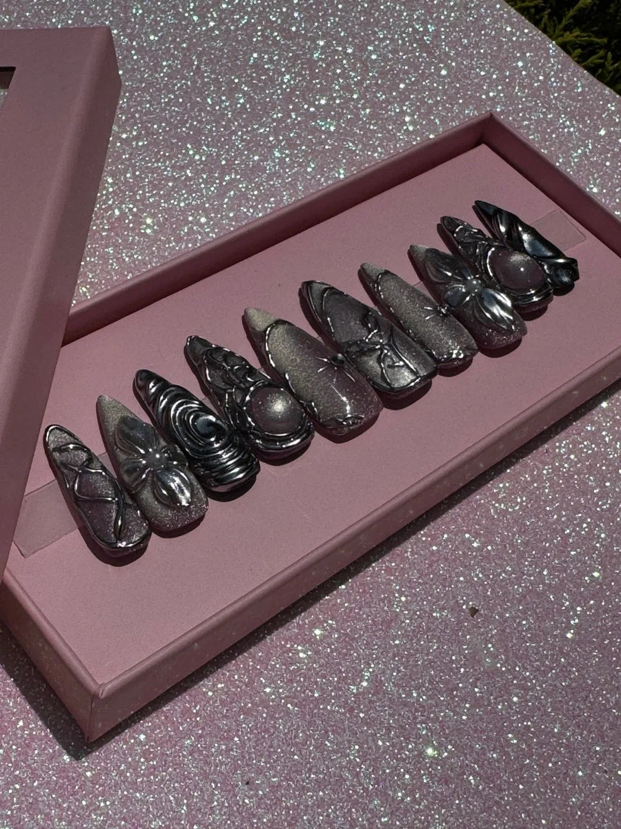 Medium-length chrome press-on nails featuring 3D silver charms and unique swirling metallic designs. Some nails have central pink stones and glitter accents.