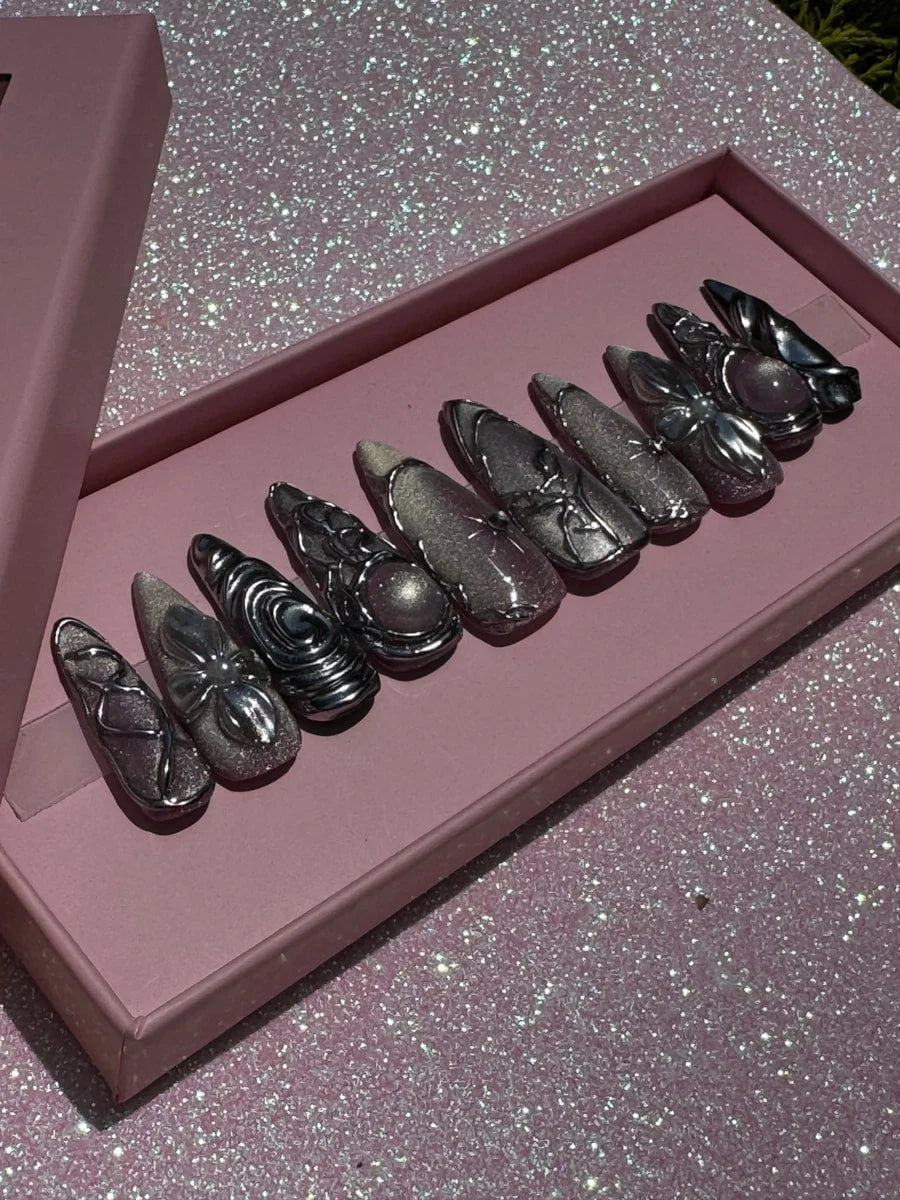Medium chrome press-on nails featuring glitter and ornate 3D silver decorations with flower, swirl, and orb accents; some nails include a central pink stone.