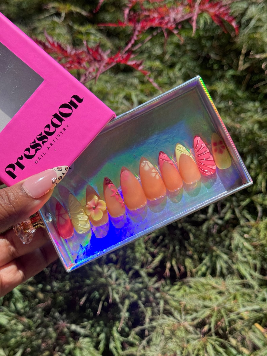 Short almond-shaped press-on nails with a springtime floral design in orange, pink, and yellow. Some nails have 3D flower accents or french tips.