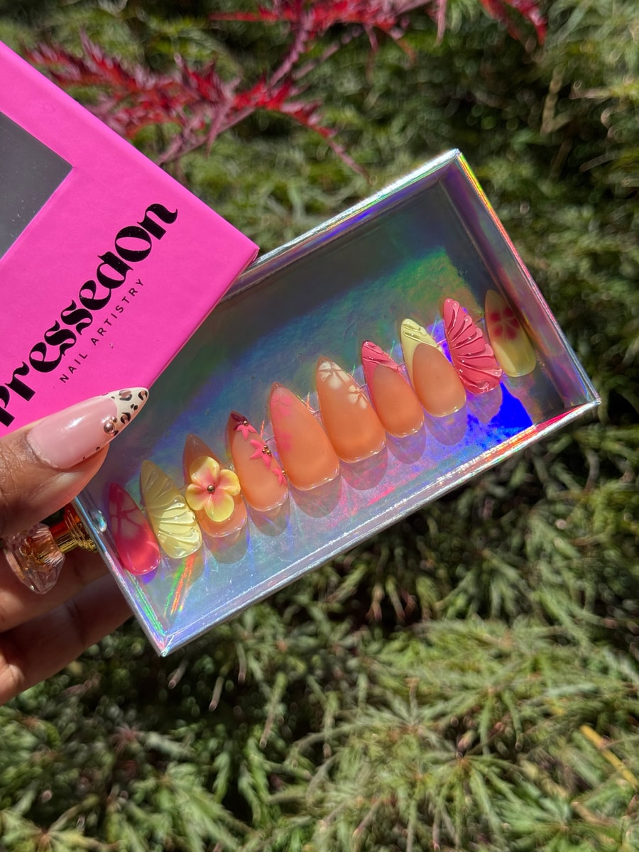 Set of almond-shaped press-on nails in orange and yellow tones featuring floral and butterfly designs. Some nails have 3D flower accents.