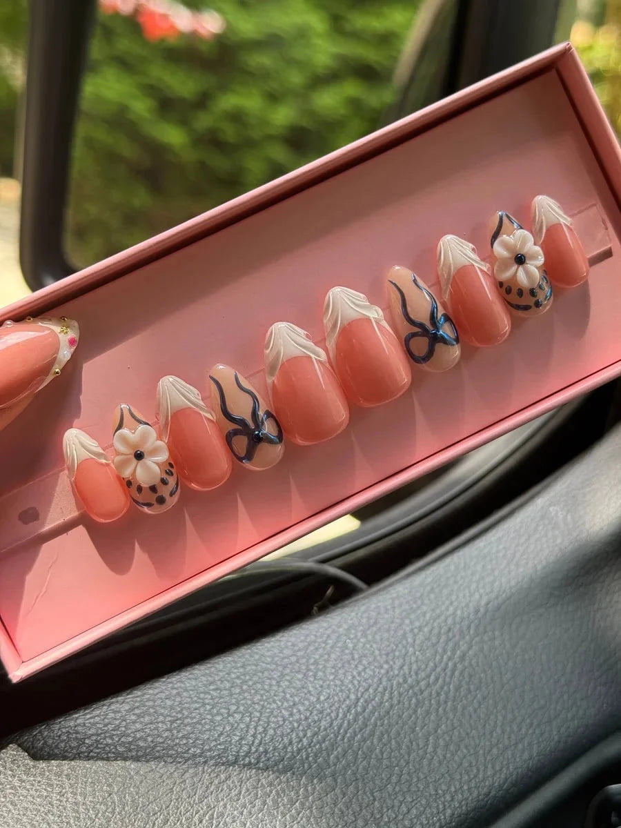 Peach-toned, super short press-on nails featuring white French tips, navy blue ribbon bows, and a 3D flower nail charm. These nails have a chic and feminine design.