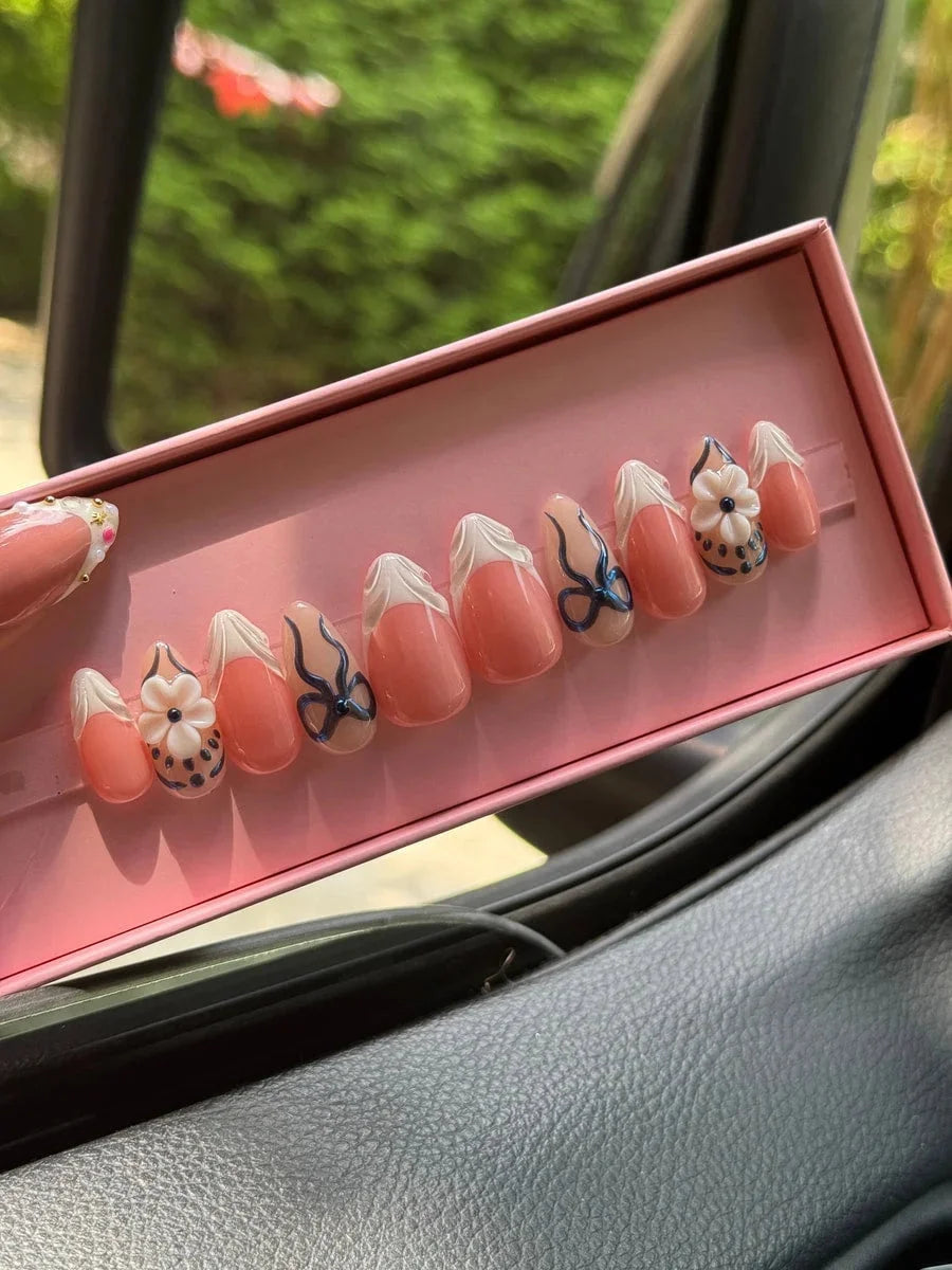 Super short peach press-on nails with white French tips, delicate blue bows, and 3D flower nail charms.