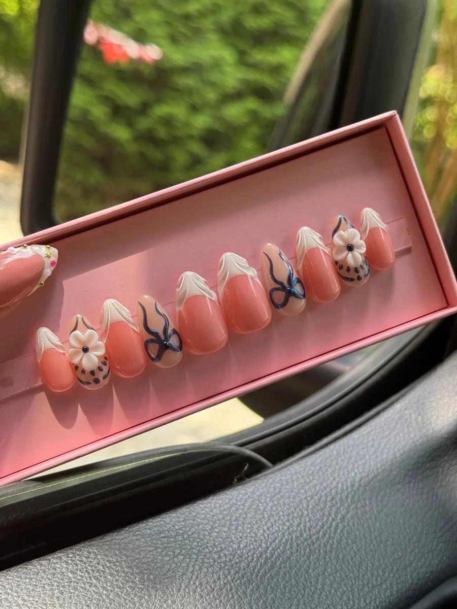 Super short peach press-on nails with white French tips, delicate blue bows, and 3D flower nail charms.