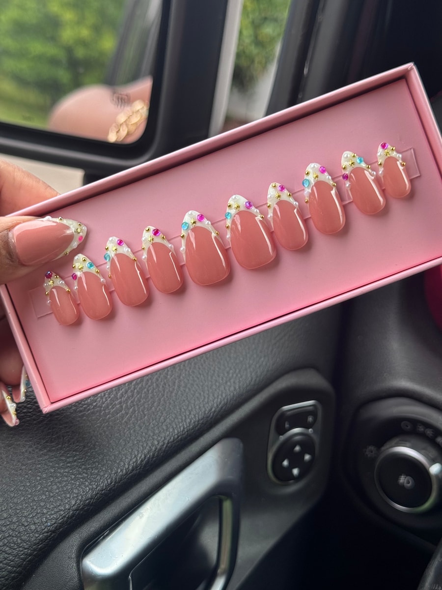 Pink-nude short press-on nails with a white French tip, decorated with small pearls, gold stars, and colorful gems.