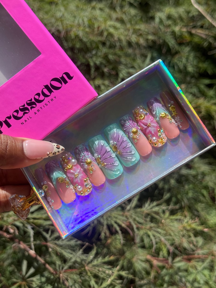 Medium press-on nails with a whimsical design in pastel colors, including pink, lavender, and light blue. The nails are decorated with floral accents, pearls, gold shells, and crystal embellishments.