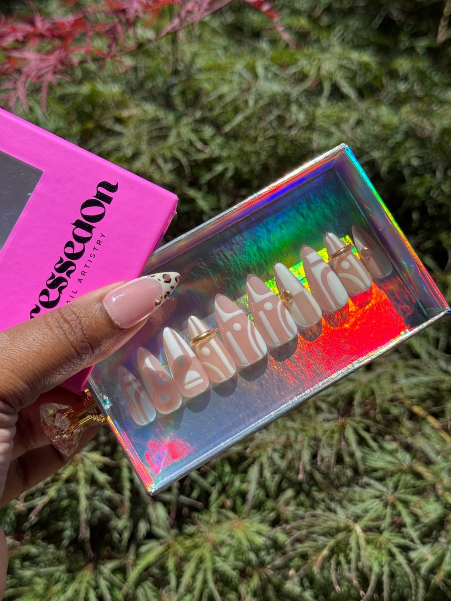 Short press-on nails with a nude base are decorated with white geometric patterns. A few nails have a single gold stud accent.