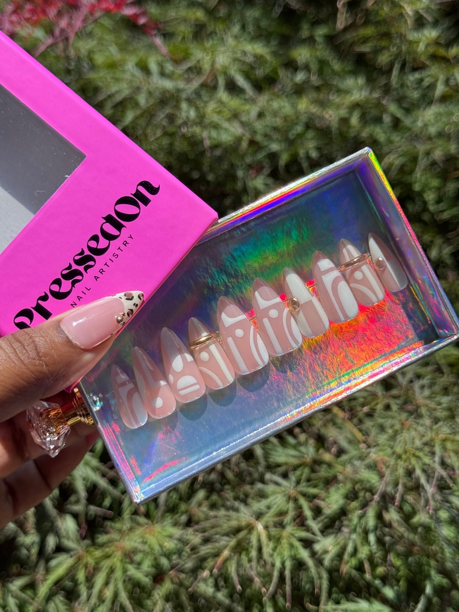 Short press-on nails display a nude base with unique white geometric designs and small gold embellishments on some nails.