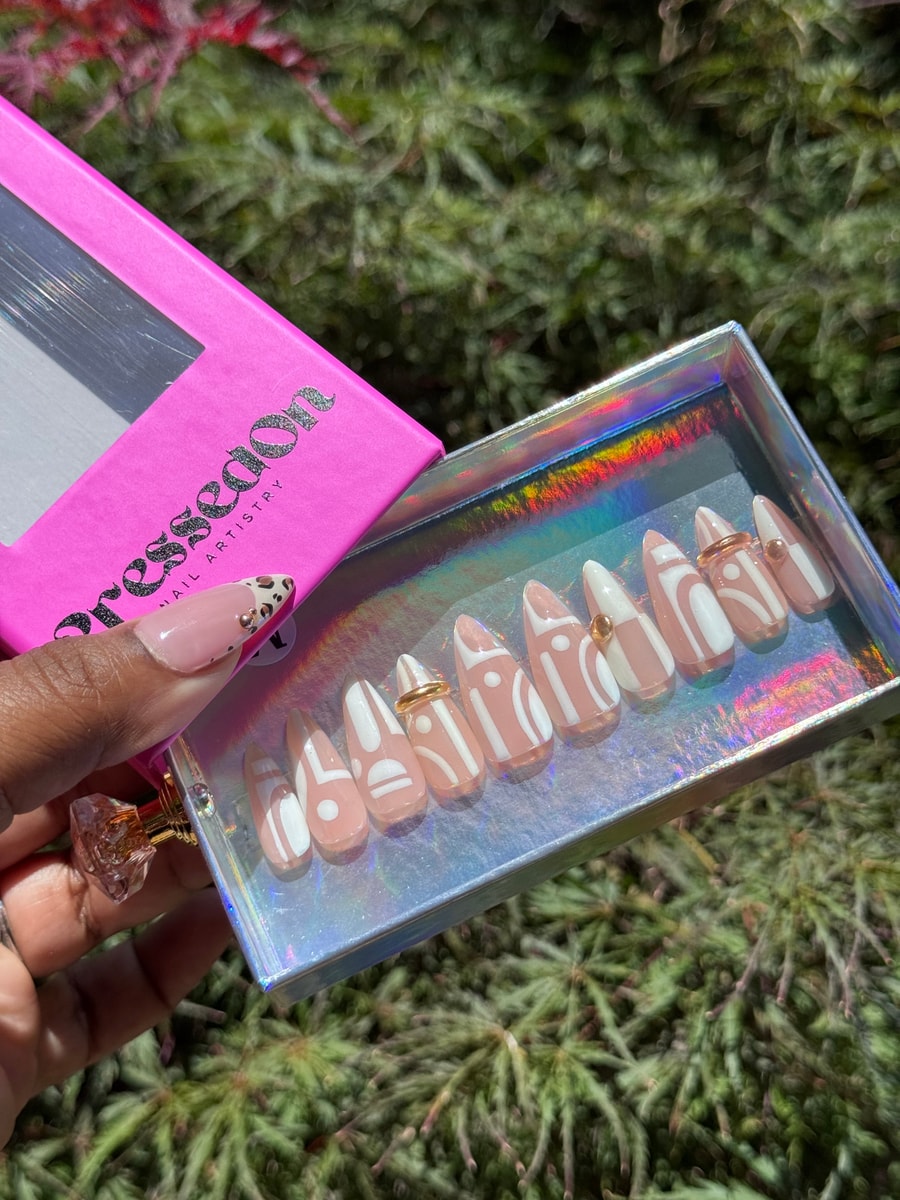 Short, nude press-on nails with white geometric patterns. Some nails feature gold bead and ring accents.