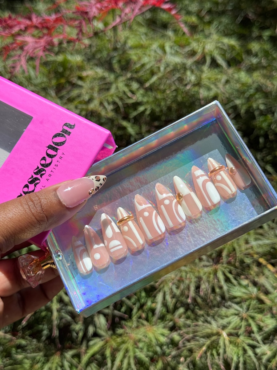 Short press-on nails with a nude base and white geometric patterns. Several nails have gold bead accents, and one includes a gold ring charm.