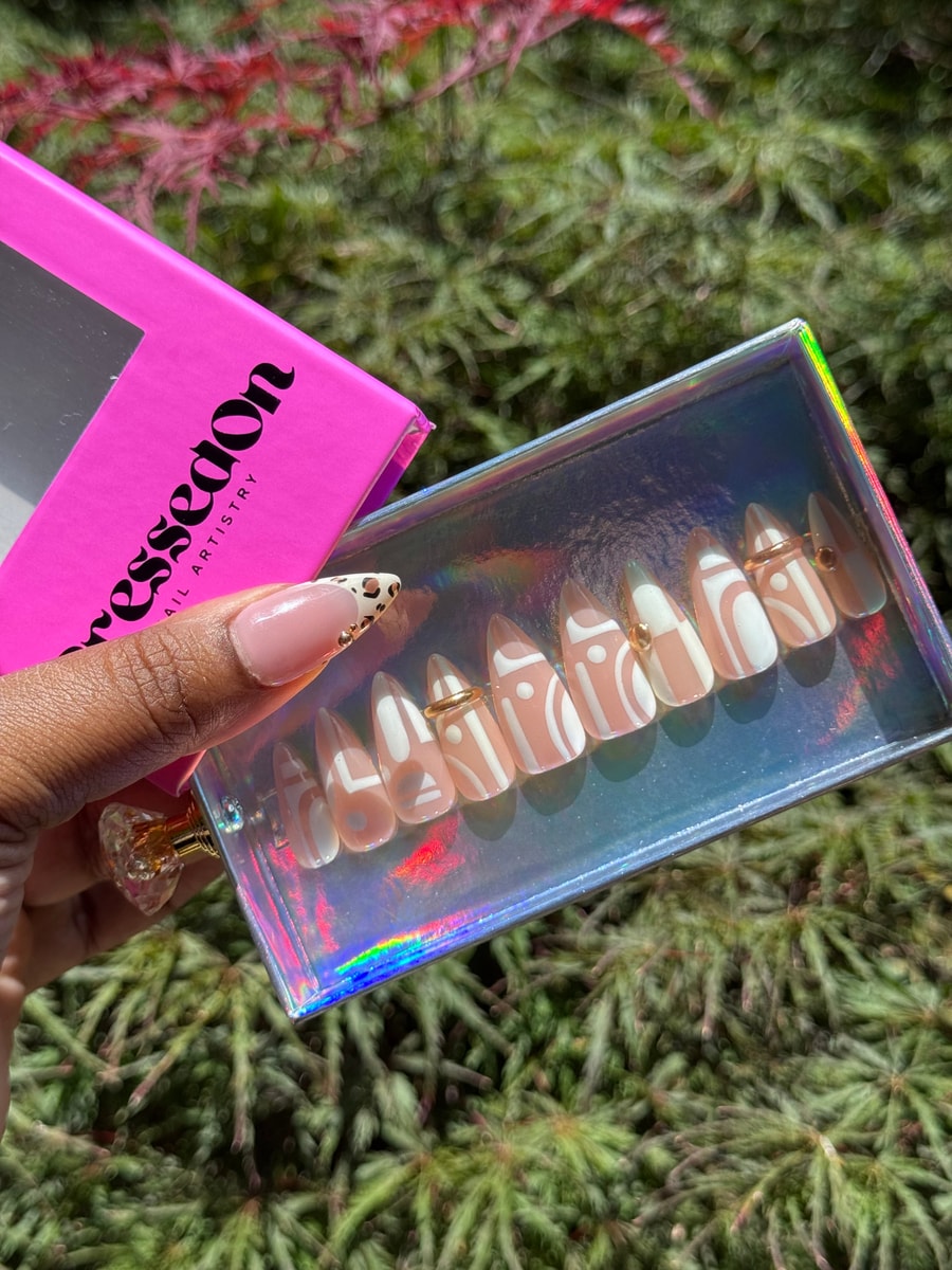 These are short, stiletto-shaped press-on nails with a nude base and white geometric designs. Each nail features a small gold bead accent. 