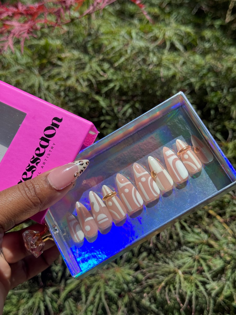 Short press-on nails with light pink base color and white geometric designs. Some nails are accented with small gold studs.