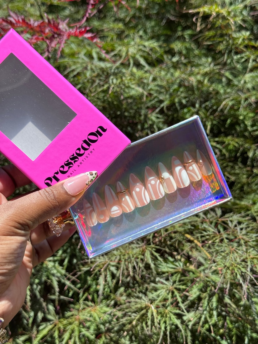 Short almond-shaped press-on nails with a nude base and white geometric designs, and gold bead accents on some nails.