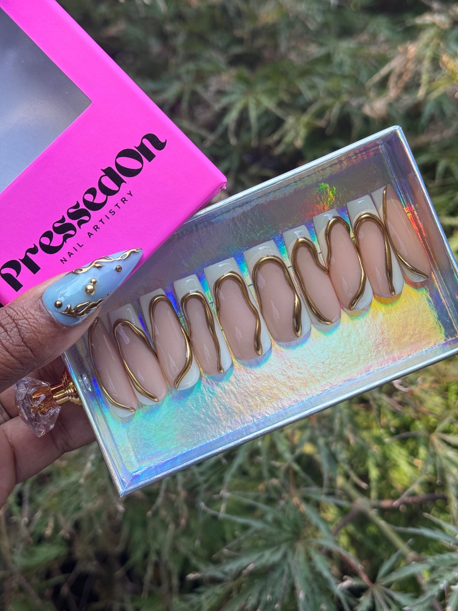 These are short, white press-on nails with nude centers and abstract gold line designs. One nail has a baby blue color with gold details.