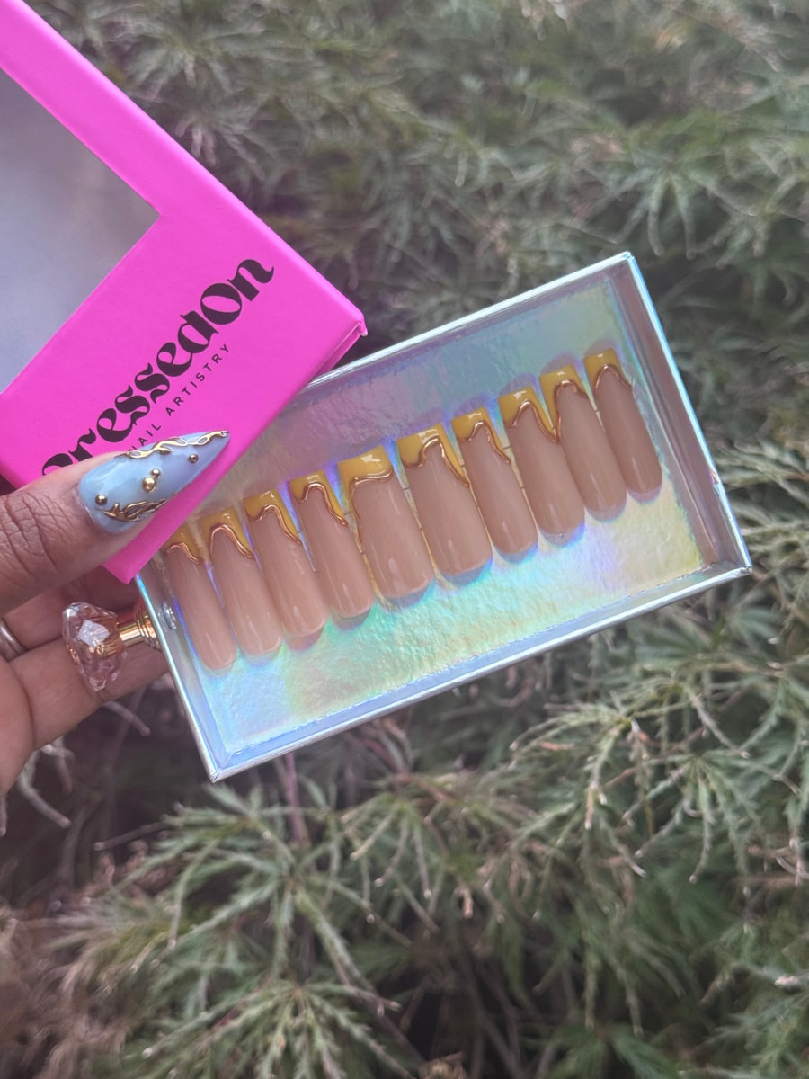 Set of medium-length press-on nails with a nude base and yellow tips with gold drip accents.