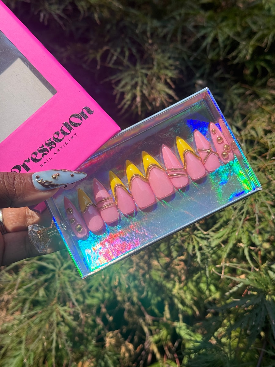 Short pink press-on nails with yellow tips and gold accents. One nail is decorated with gold studs.