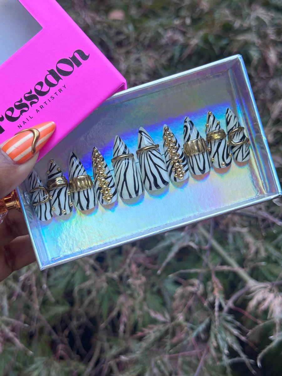 Stiletto-shaped press-on nails with a striking zebra print, adorned with gold rings and small gold studs, creating a unique and fashionable look.